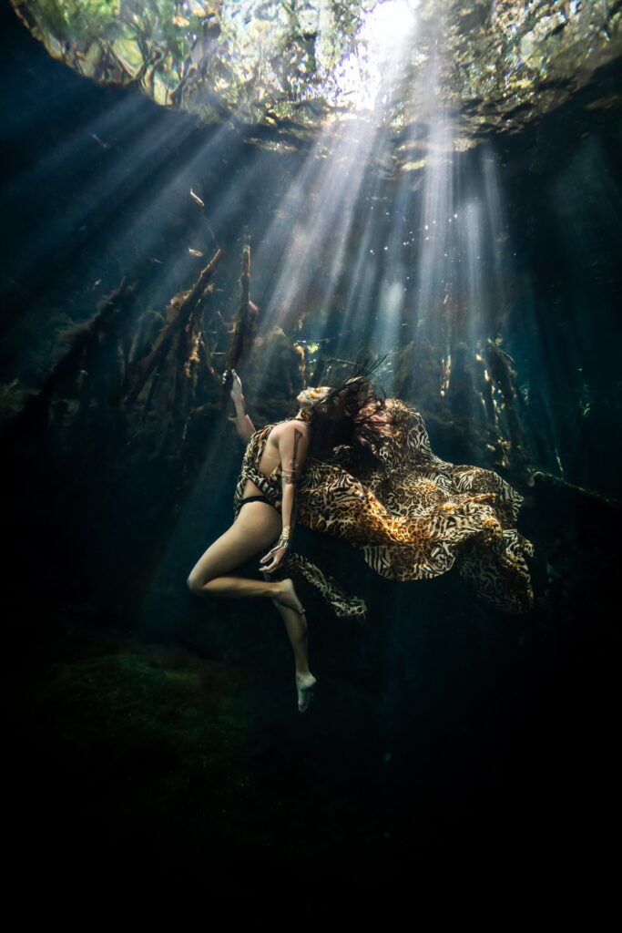 Artistic underwater portrait of a woman wearing a leopard-print dress, with skull-inspired makeup and a decorative headdress, reflected on the surface of a cenote.